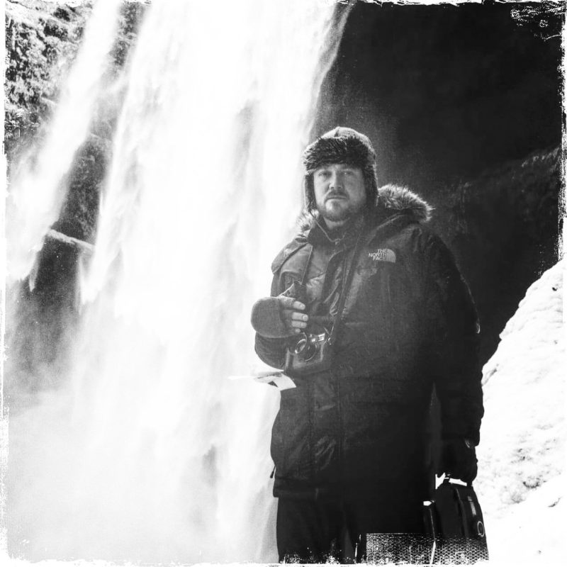 David Gensler standing near a waterfall in winter, wearing The North Face outerwear and holding a camera in a natural landscape