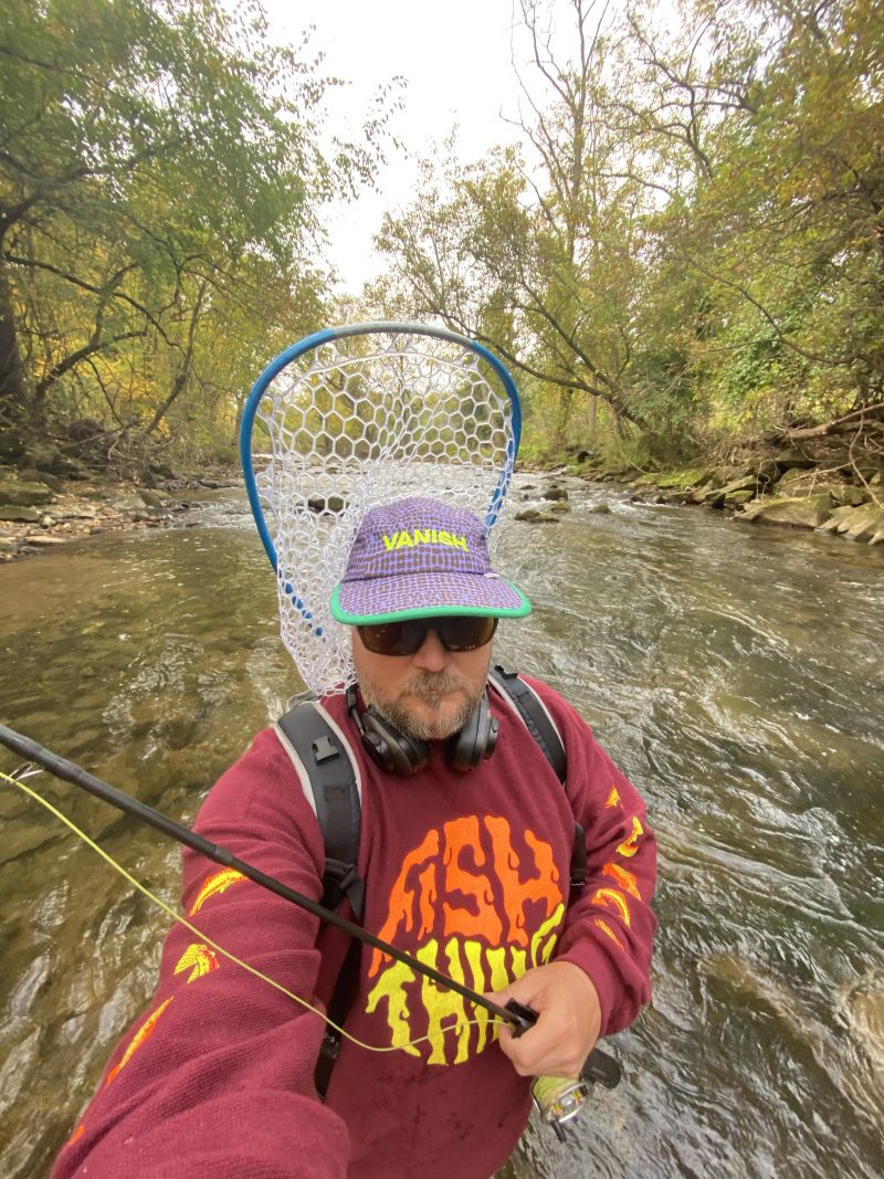 David Gensler standing in a creek wearing a Fish Thing sweatshirt and Vanish hat while holding a fishing rod.