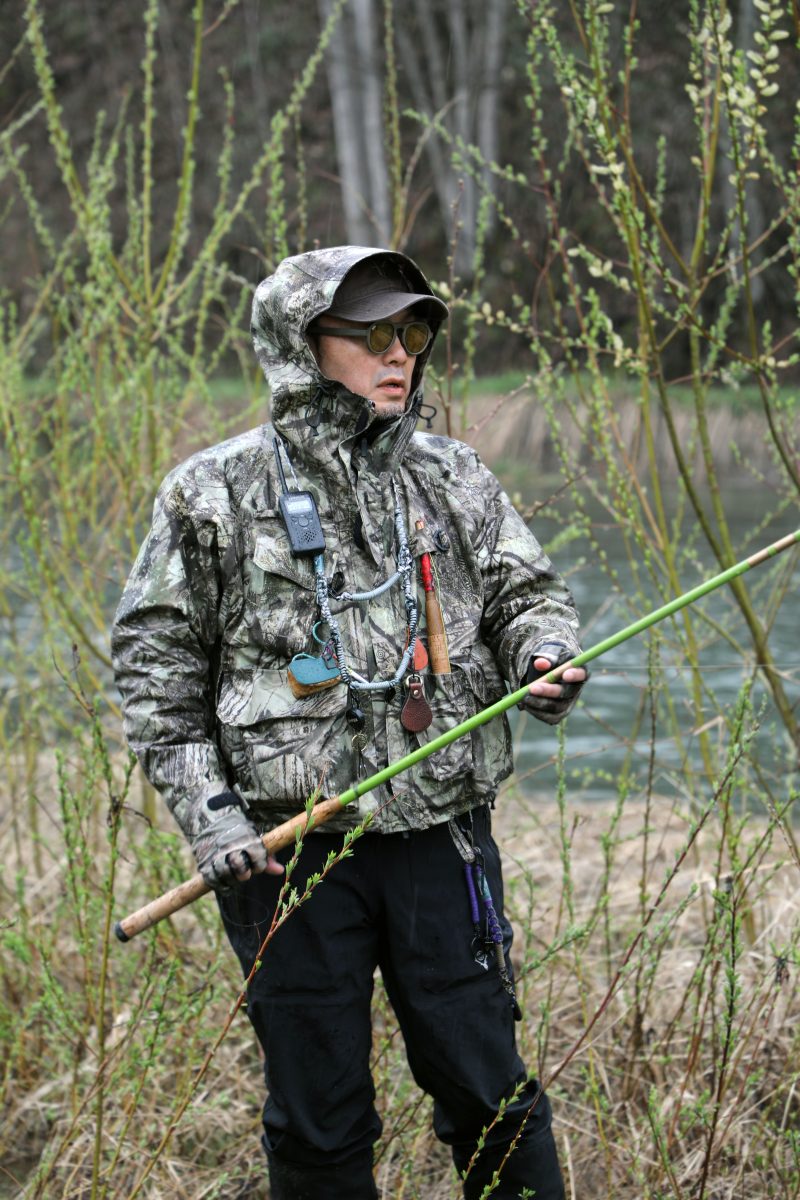 David Gensler outdoor style reference showing a South2 West8 camouflage fishing jacket worn in a riverside setting with a fishing rod.