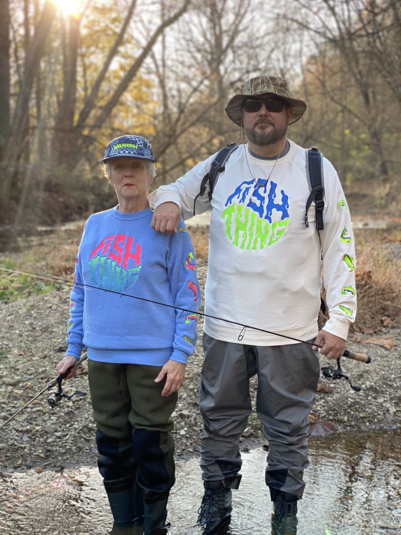 Sharon and David Gensler standing in a shallow creek wearing Fish Thing sweatshirts and fishing gear.
